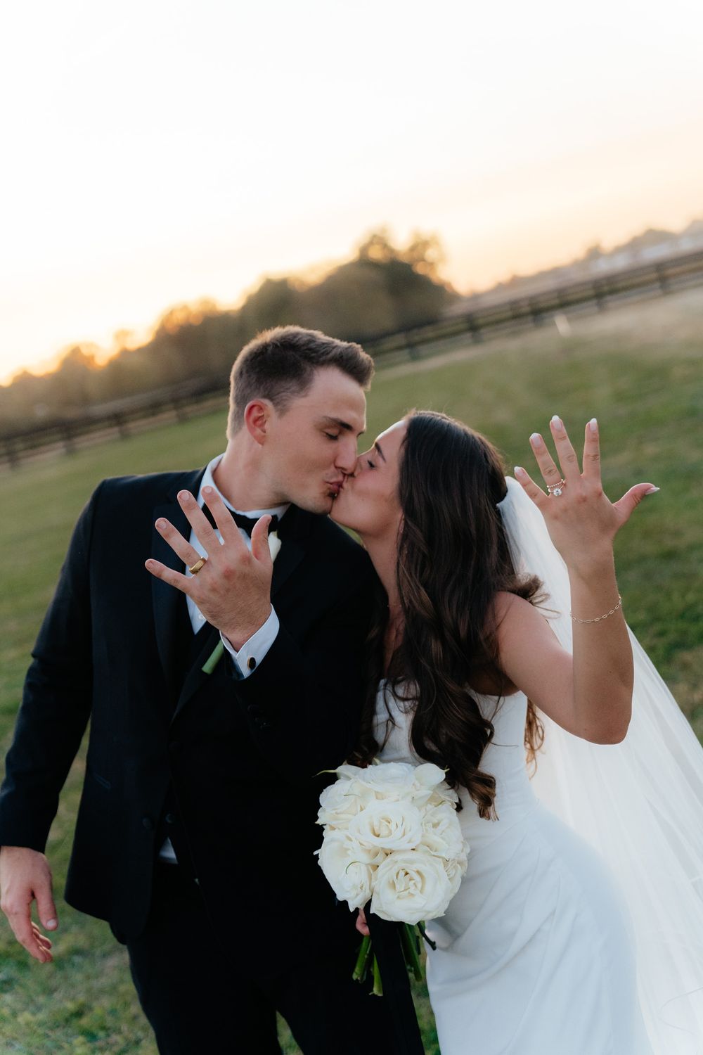 Couple in wedding attire showing rings while kissing at sunset.