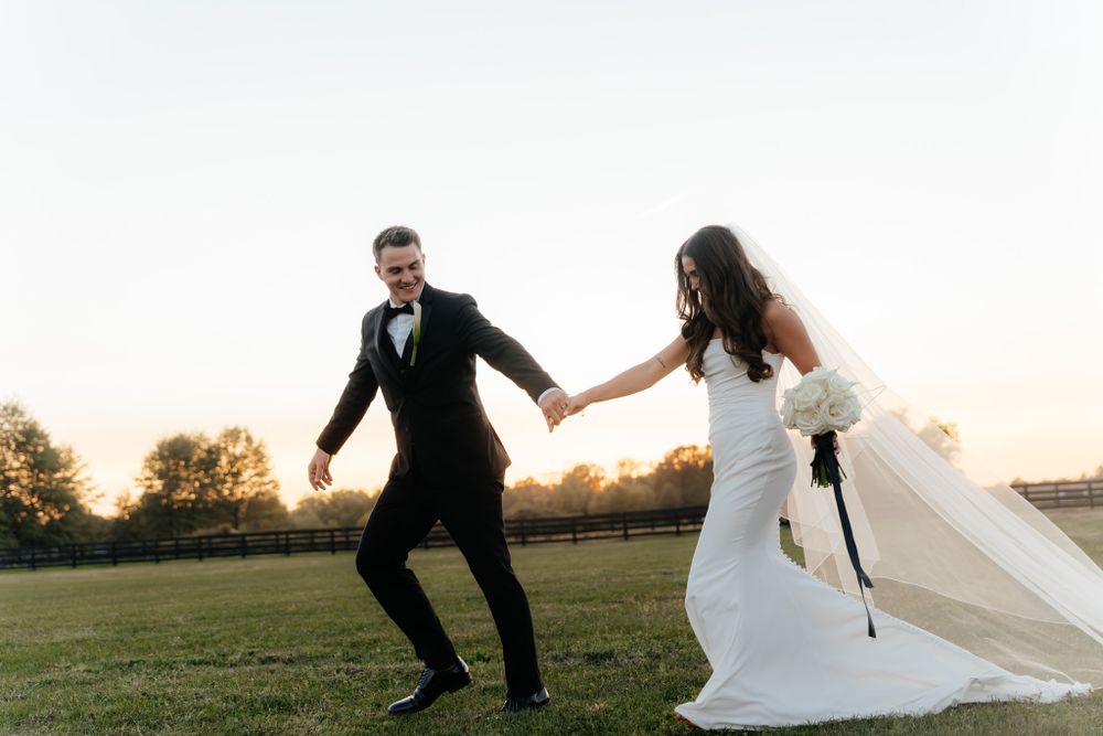 A bride and groom run joyfully across a grassy field at sunset, her white veil flowing in the breeze.