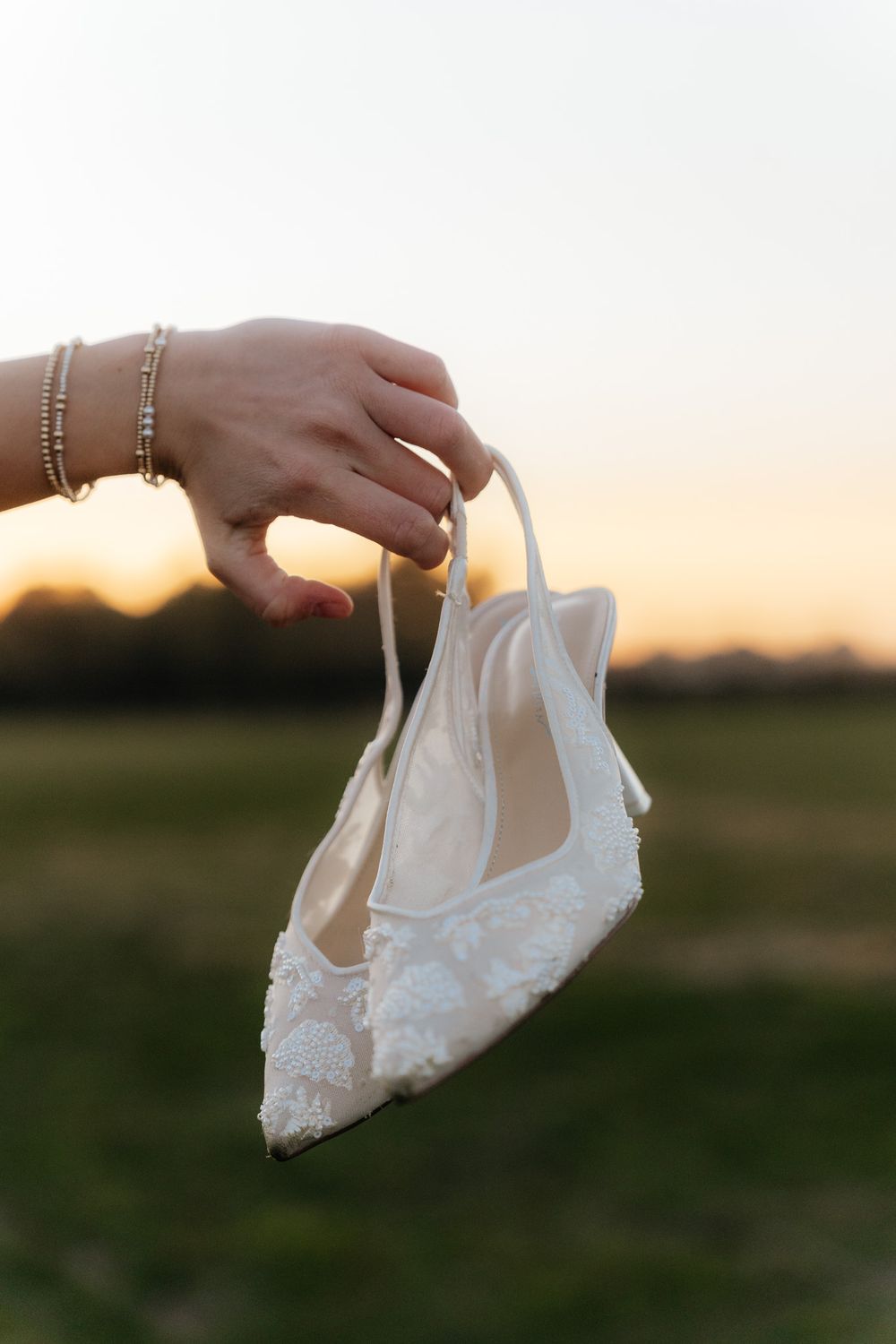 A hand holding delicate white lace bridal shoes against a sunset backdrop.
