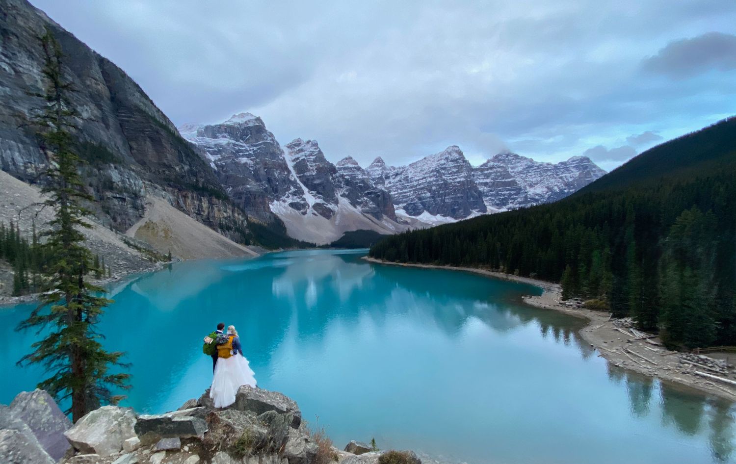 Wedding couple admiring the turquoise waters of Moraine Lake with snowy mountain peaks in Banff National Park. Banff elopement photography