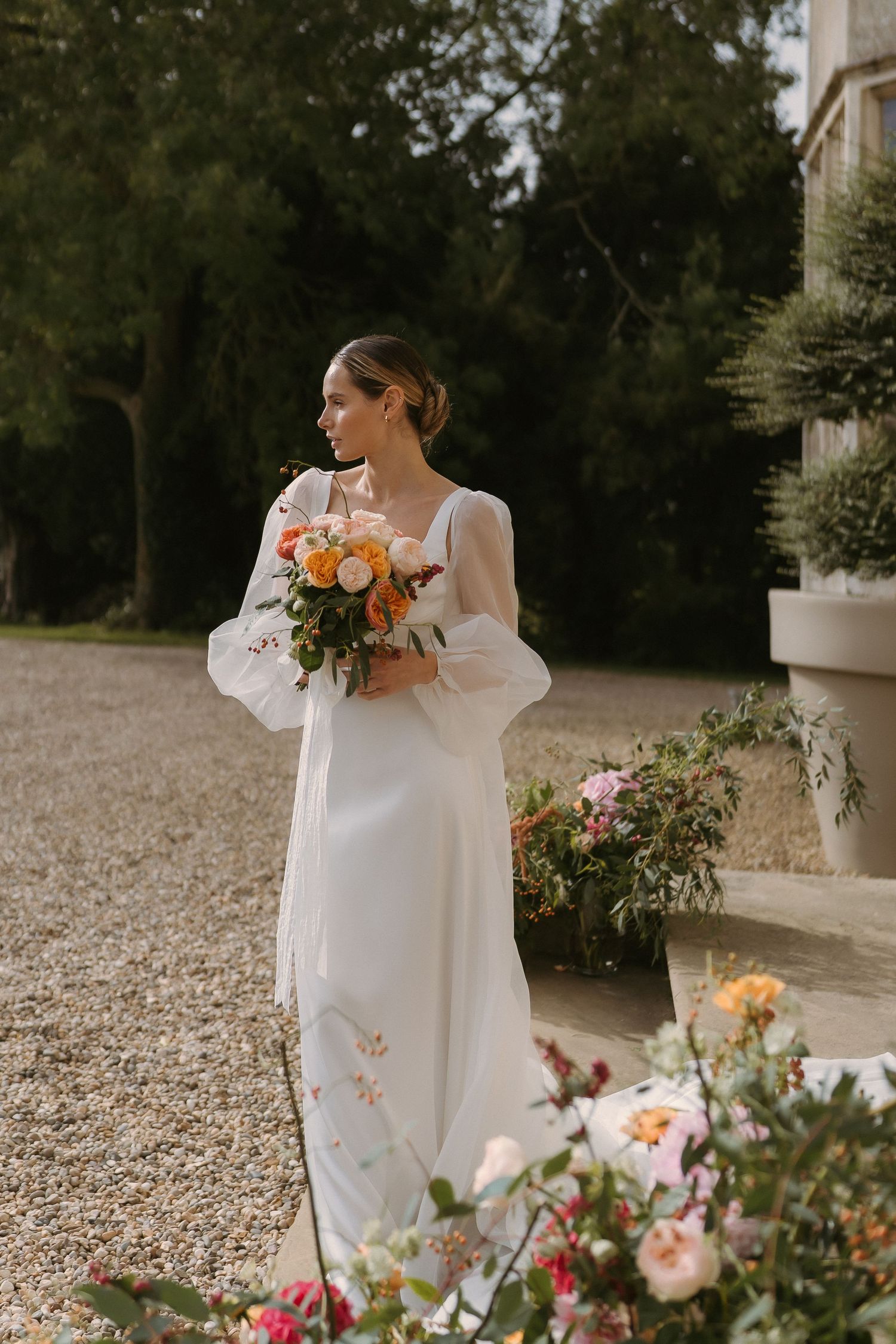 An elegant bride in a white romantic Jesus Peiro gown holds a peach and orange bouquet on the steps of Howsham Hall.