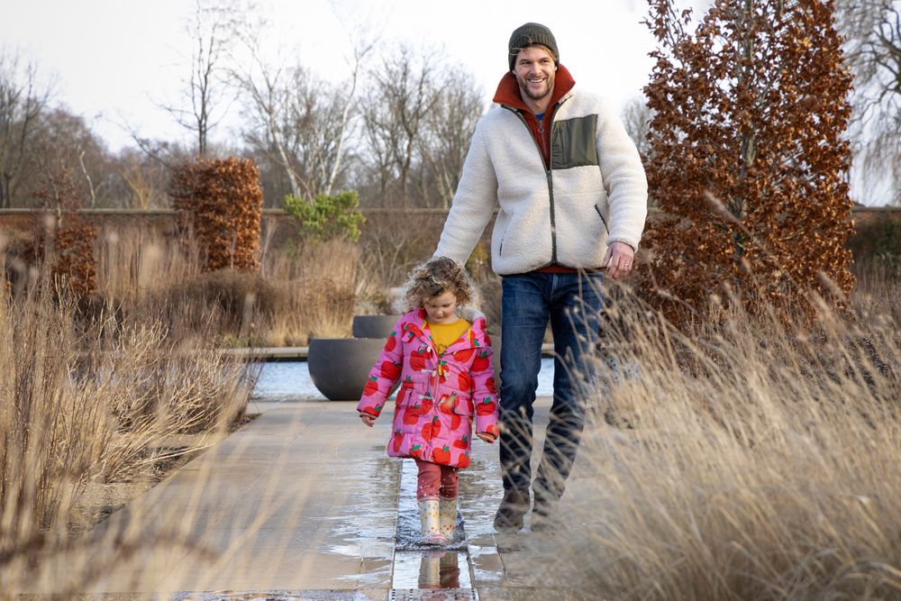 Two people walking along a wooden boardwalk path through tall winter grasses on an overcast day.