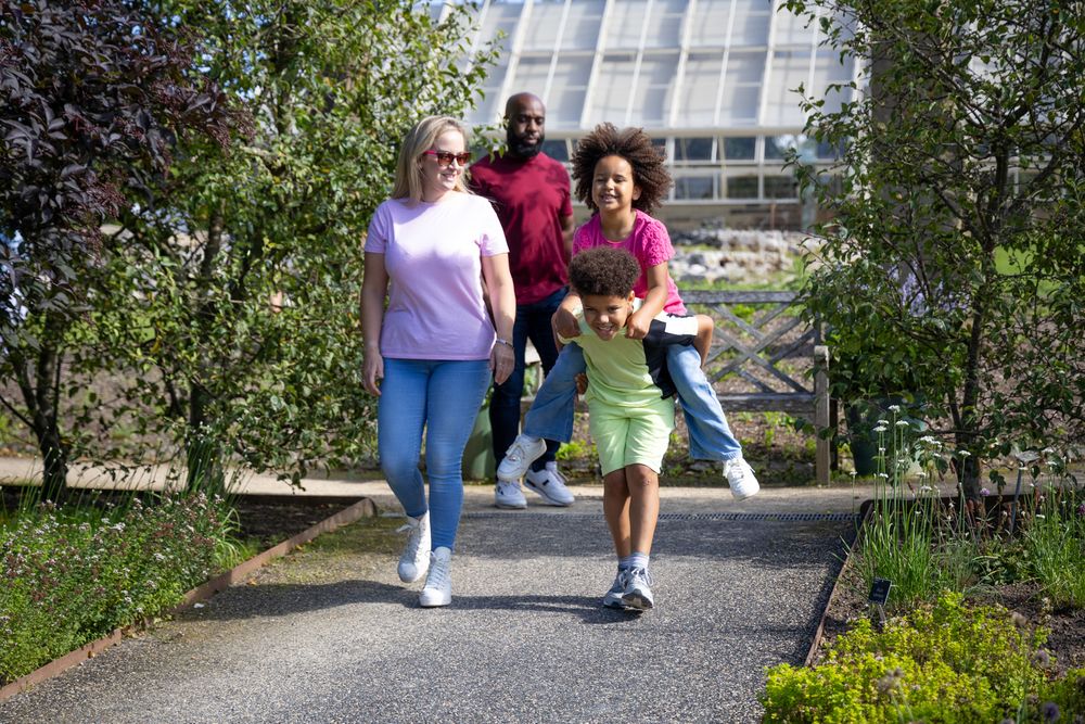 Three people stroll together on a winding garden path with greenery and a modern building in the background.