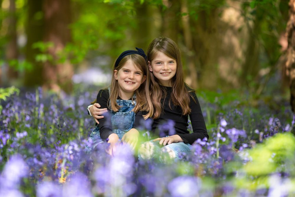 Two people sit together in a spring bluebell woodland surrounded by vibrant purple flowers in dappled sunlight.