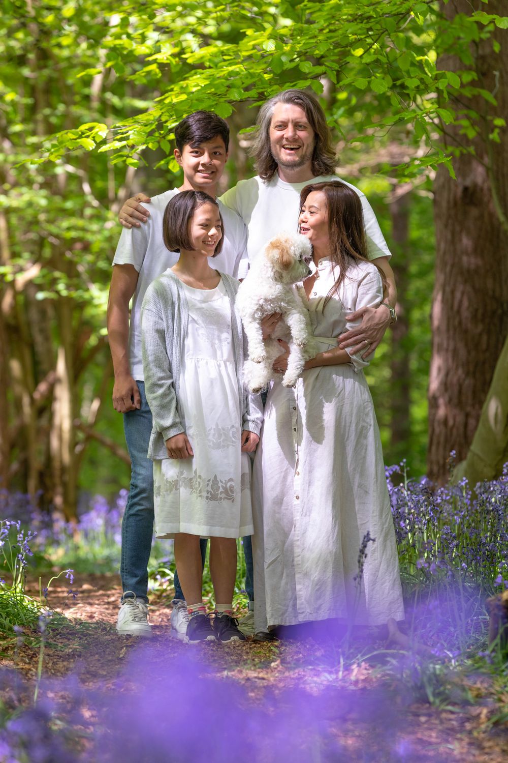 A family stands together on a woodland path with their small white dog surrounded by blooming purple wildflowers.