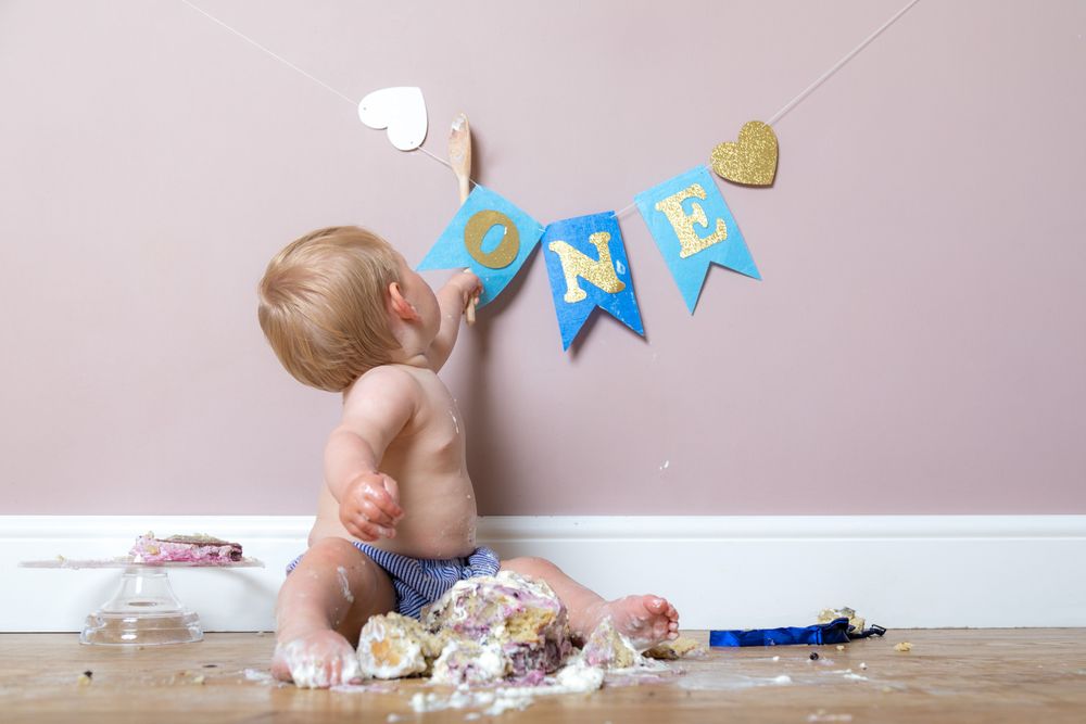 Baby in a diaper sits on floor with smashed birthday cake near blue and gold first birthday banner decorations.