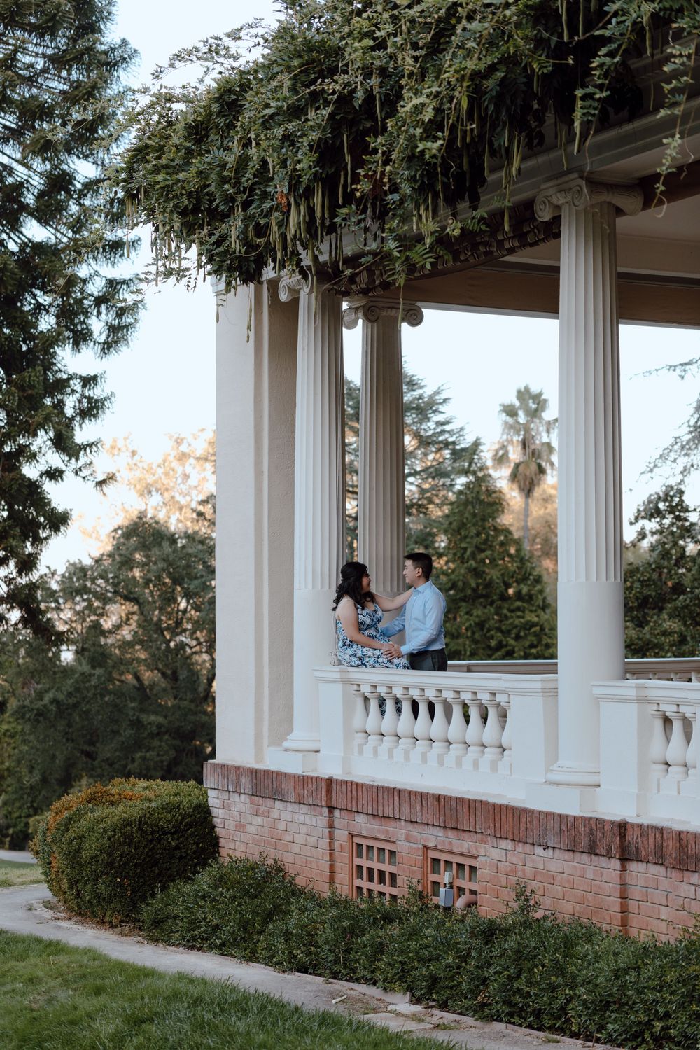 A romantic couple shares a moment on an elegant white-columned porch with greenery draping overhead in a garden setting.