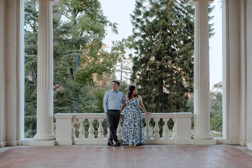 A romantic couple stands on an elegant white columned balcony surrounded by towering evergreen trees at sunset.