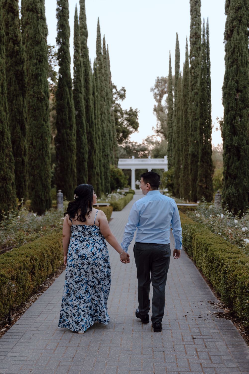 A couple walks hand in hand down a cypress tree-lined garden path with brick pavers.