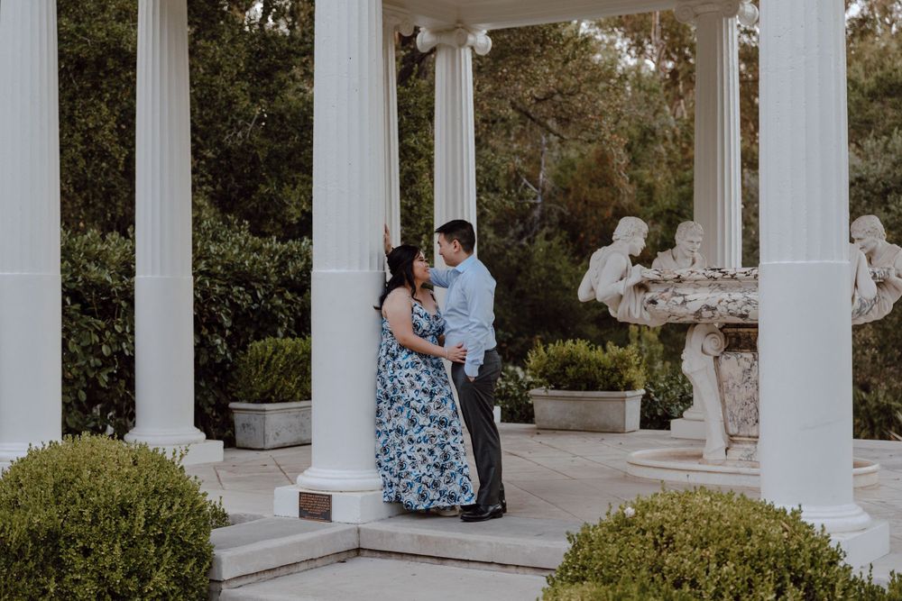 A romantic couple embraces between elegant white columns in a garden setting with classical statuary visible.