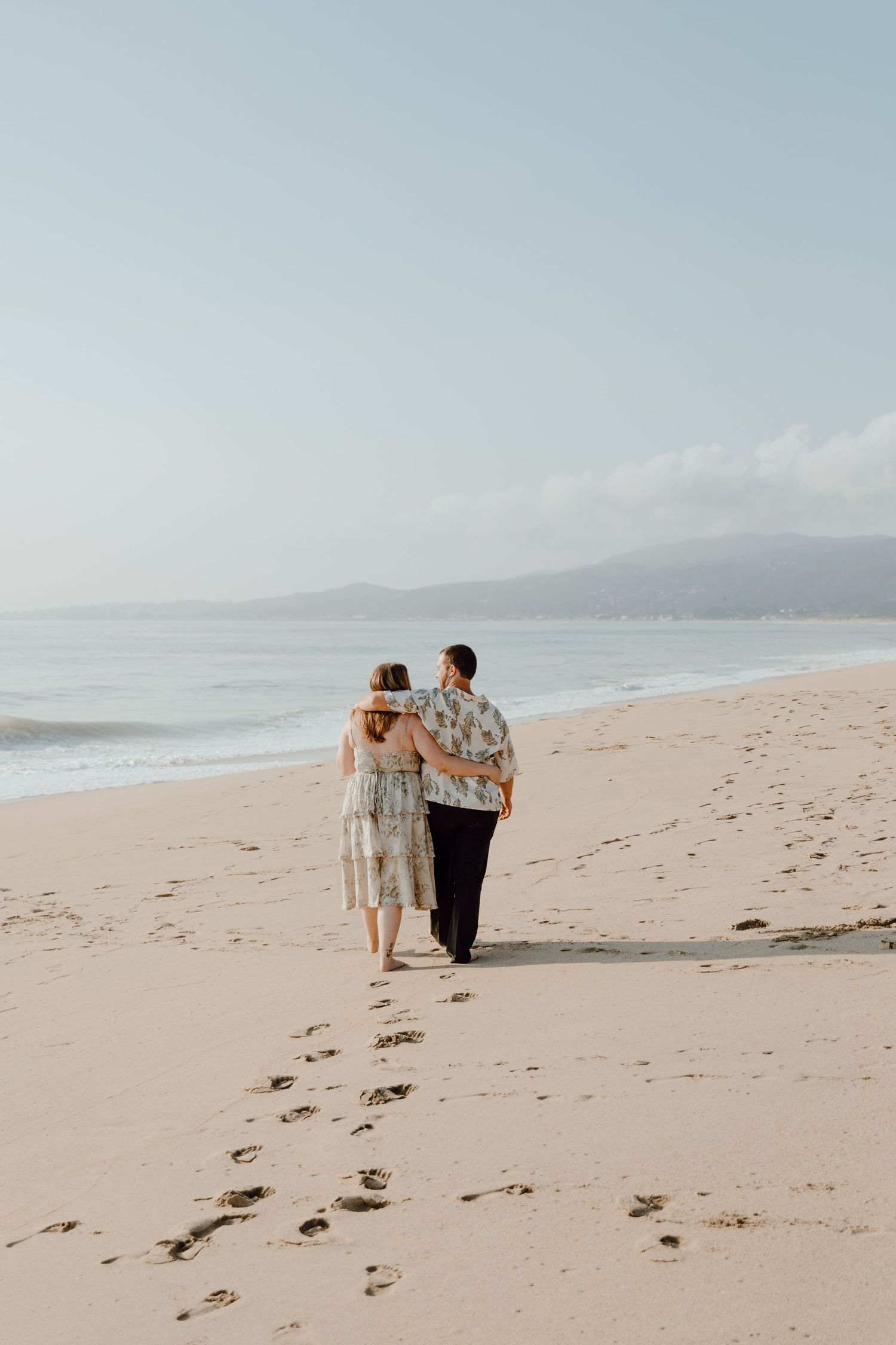 A couple walks together along a peaceful beach leaving footprints in the sand as mountains rise in the distance.
