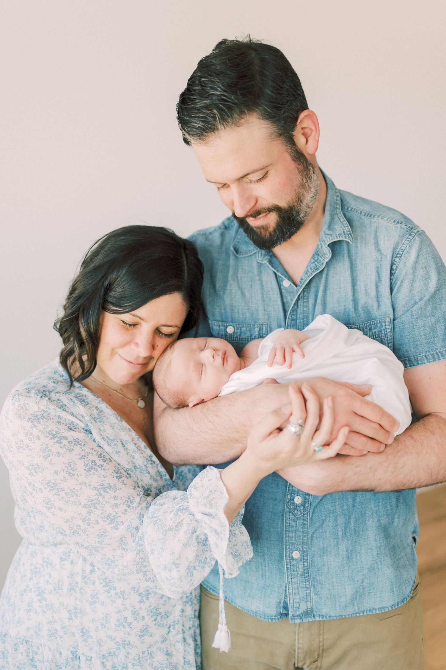 Two adults in soft neutral tones hold and gaze lovingly at a sleeping newborn baby against a light backdrop.