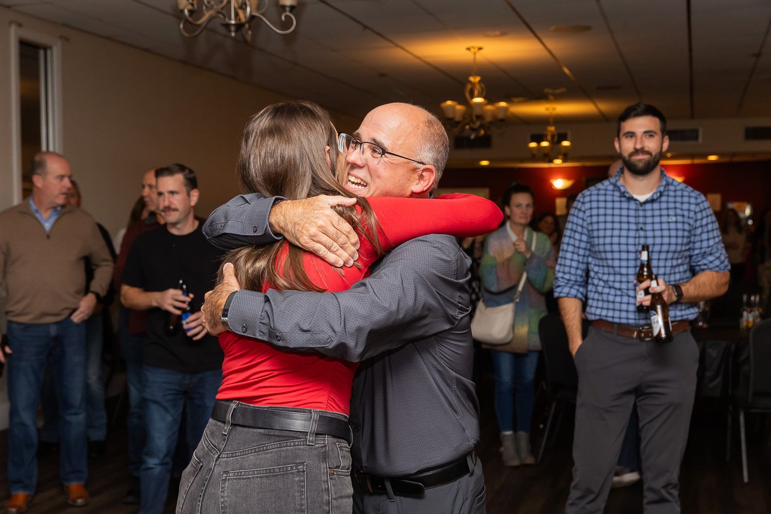 People share embraces during an indoor social gathering with chandelier lighting.