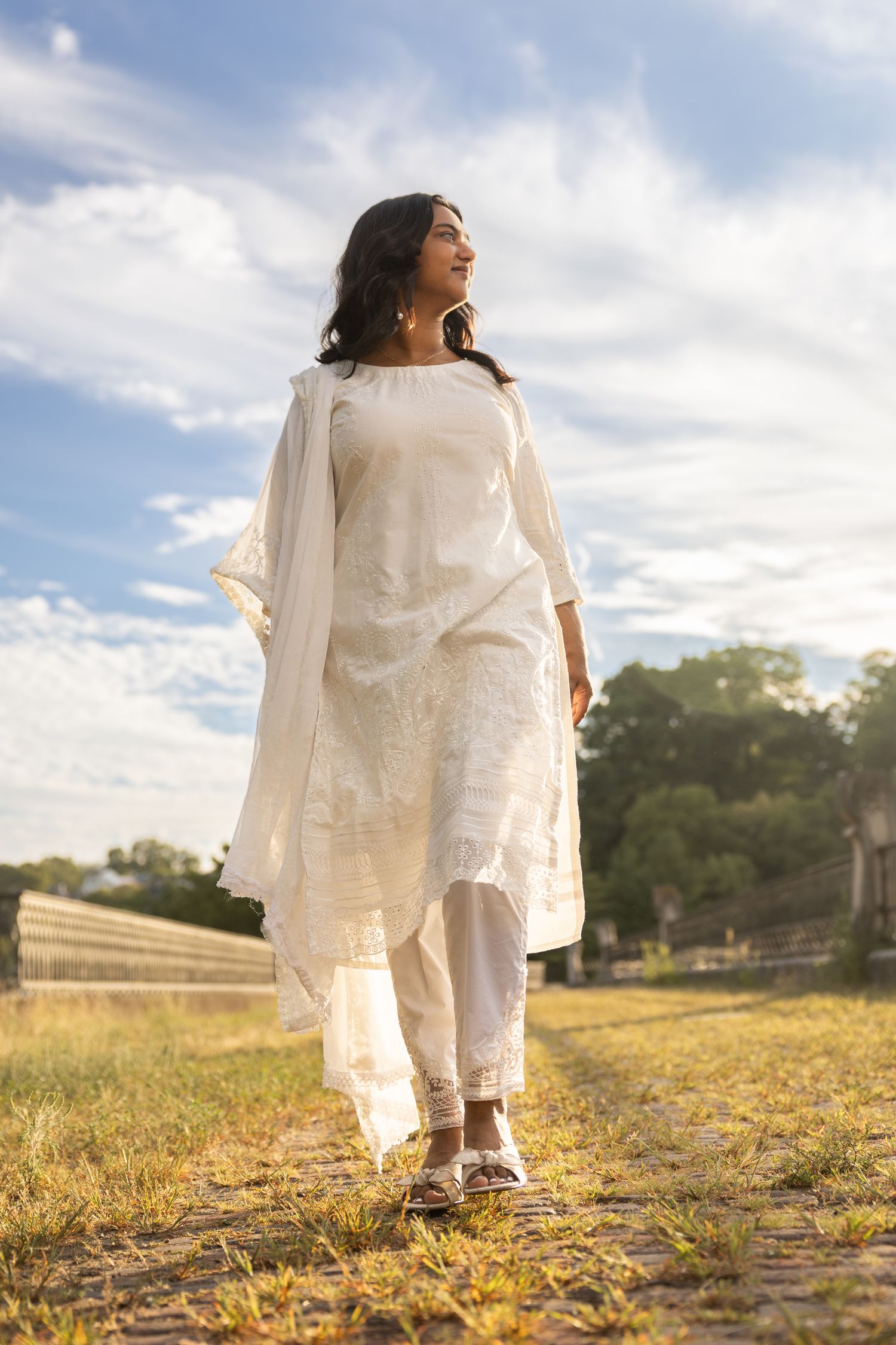 Person in flowing white traditional dress stands in golden field at sunset against cloudy blue sky.