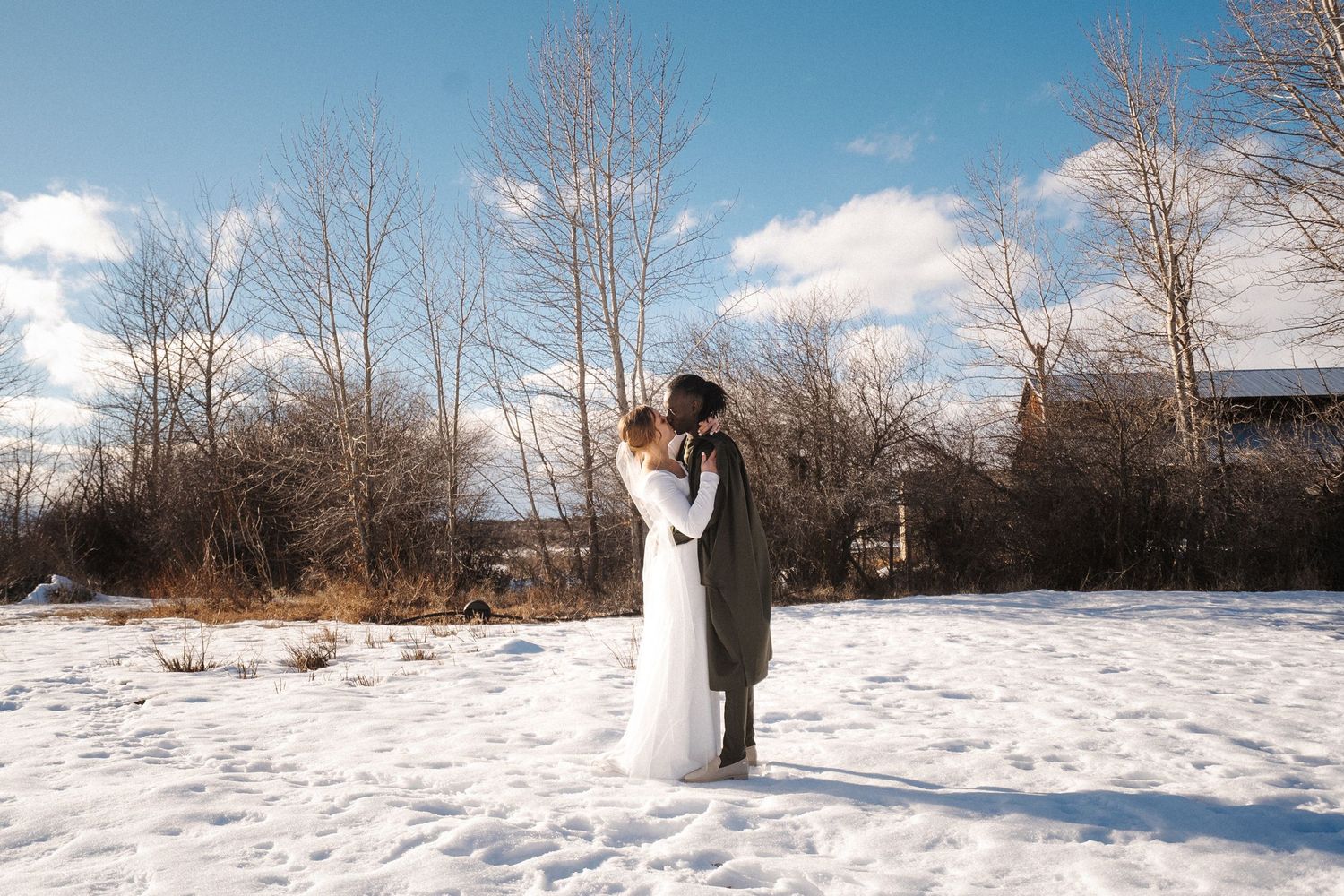 A couple shares an intimate moment while standing in a snowy field surrounded by bare winter trees and bright blue sky.