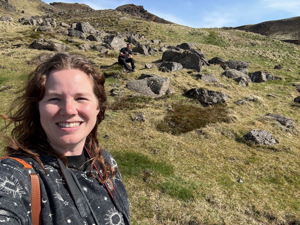 A hiker on a rocky mountain slope with scattered boulders and green grass under blue skies.