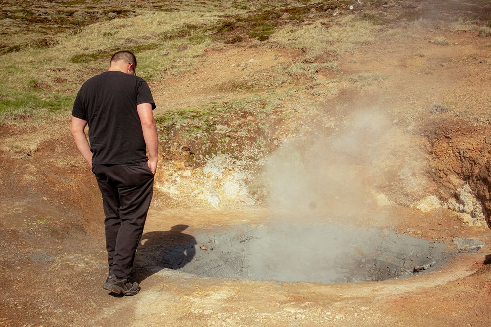 A person observes a steaming geothermal hot spring in rocky terrain beneath a brown hillside.