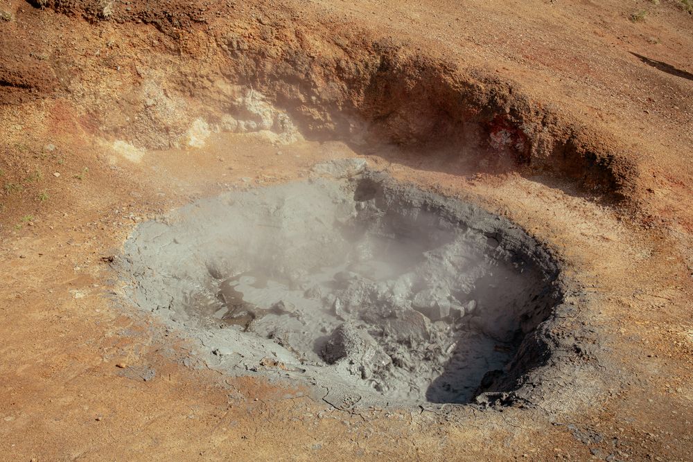 A steaming mudpot with gray bubbling mud in a reddish-brown volcanic crater formation.