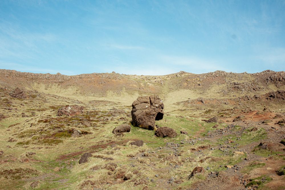 Rugged desert landscape with red-tinted rock formations and scattered boulders under bright blue sky.