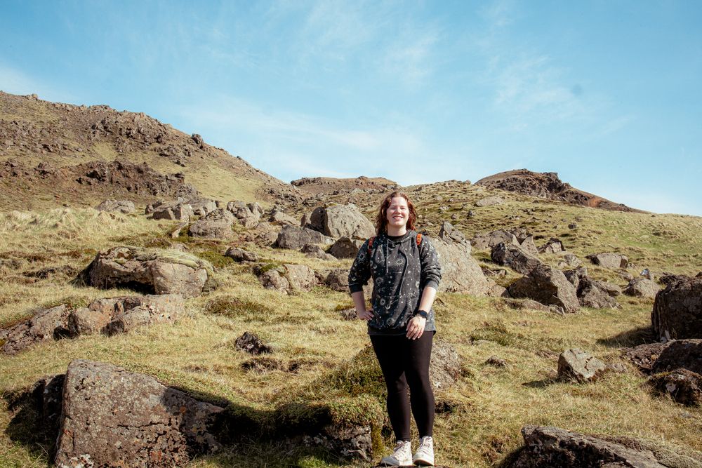 A traveler stands among scattered boulders on a grassy hillside under bright blue skies in a rugged terrain.