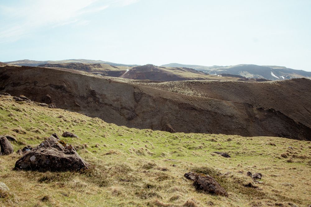 A grassy slope on a barren hillside with winding dirt trails carved into distant brown mountains under blue skies.