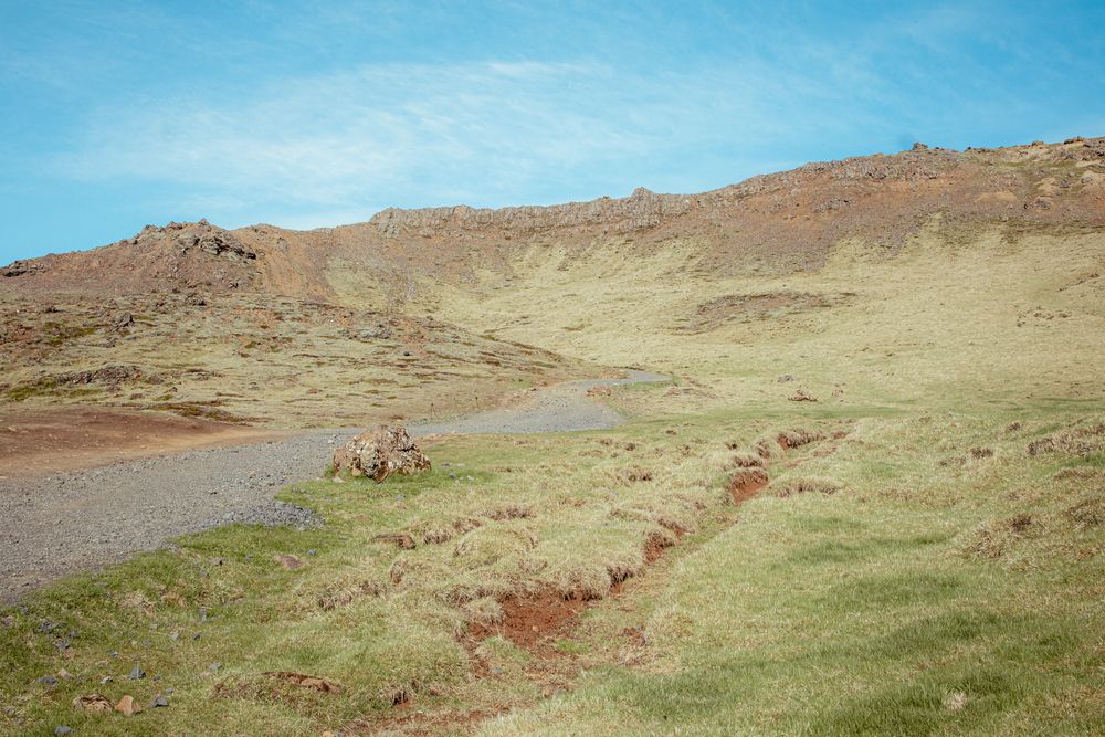 A winding dirt path leads up a barren hillside with reddish soil patches and sparse vegetation under blue skies.
