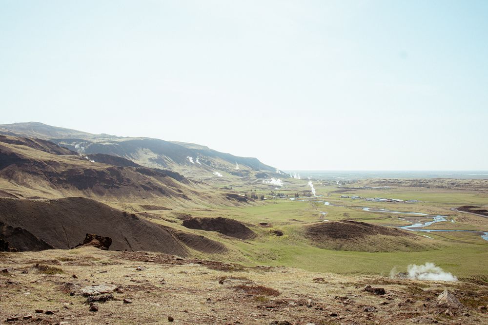 A scenic landscape view of rolling brown hills and green valleys with distant mountains under a bright blue sky.