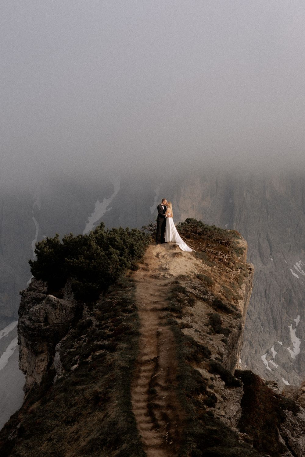 A couple stands atop a misty mountain cliff edge in dramatic wedding attire during an adventurous elopement photoshoot.
