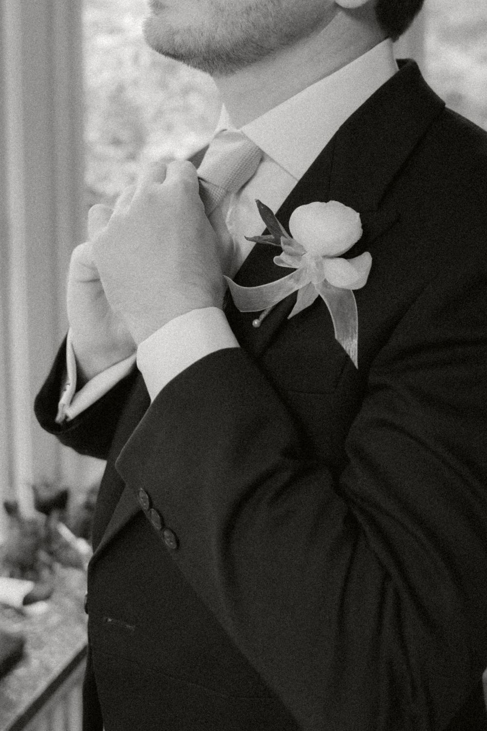 Black and white close-up of groom adjusting tie while wearing dark suit with white boutonniere flower.