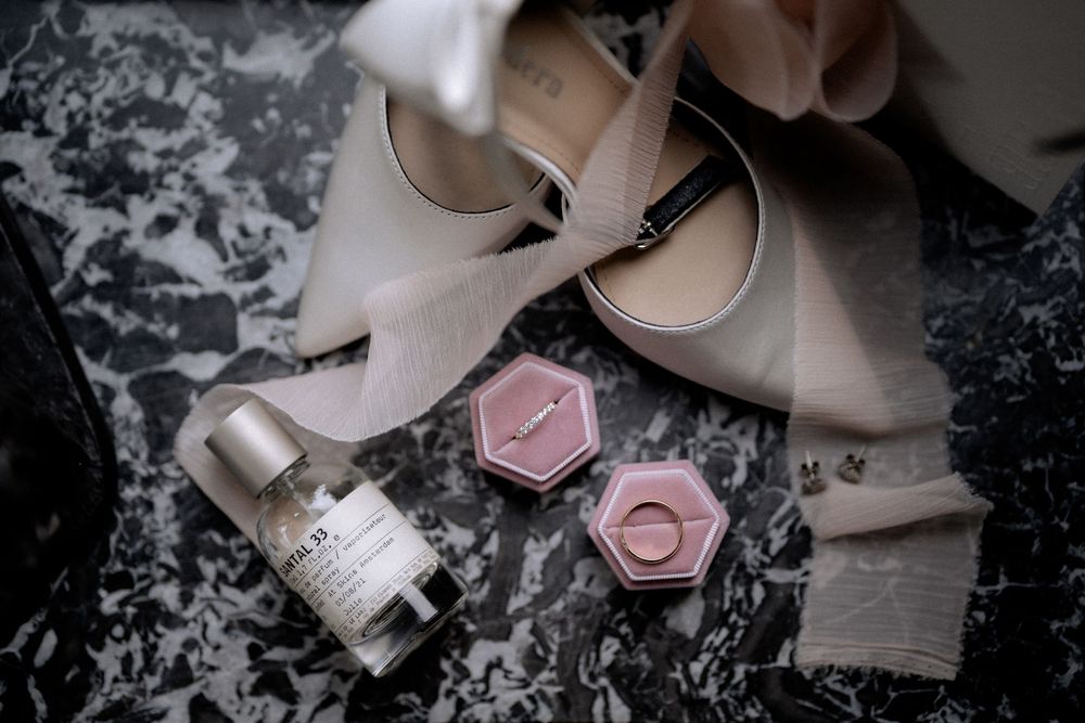 Wedding details flatlay with blush pink high heels, ring boxes and perfume bottle on marble background.