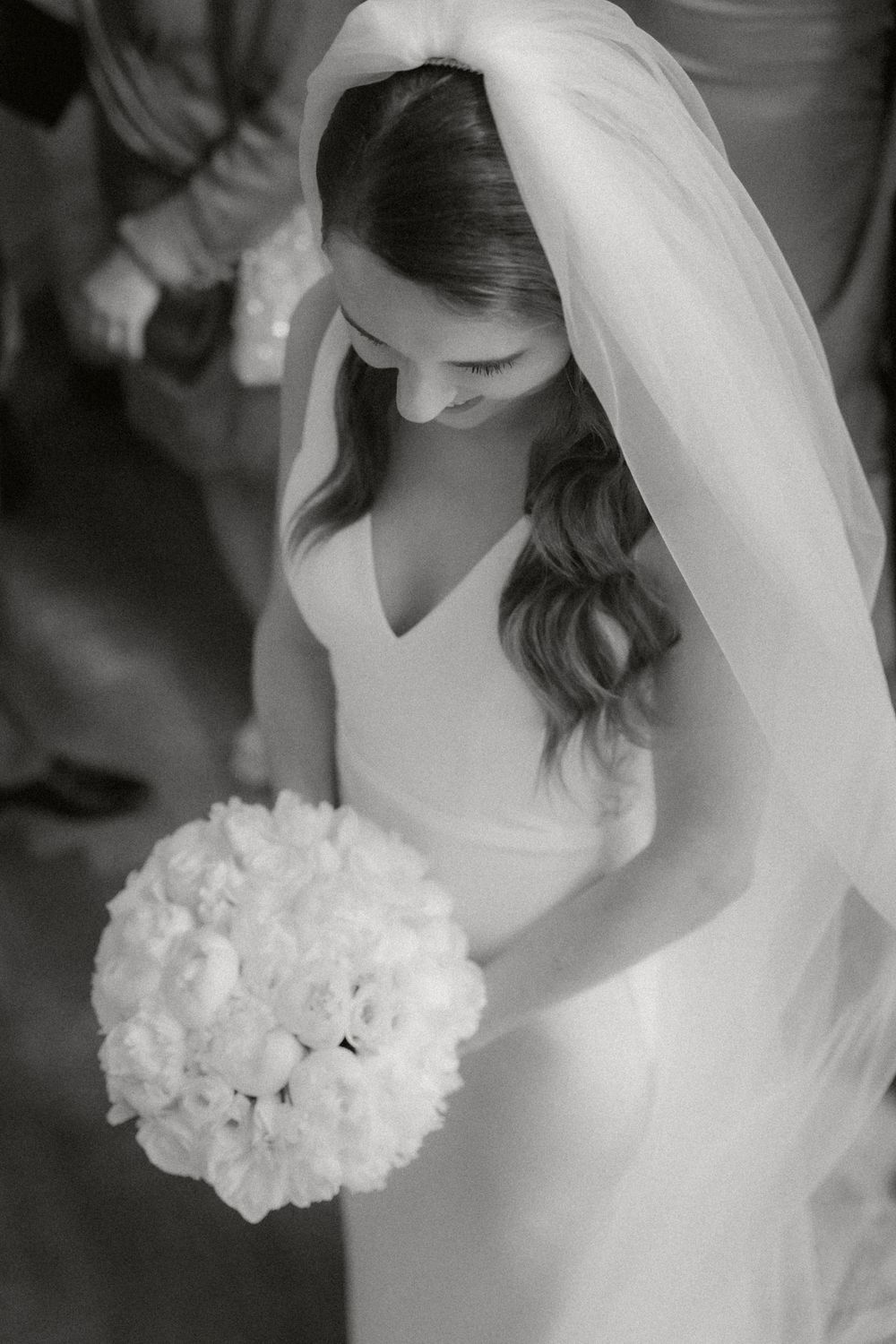 Black and white photo of bride in elegant sleeveless gown looking down at round white floral bouquet with long flowing veil.