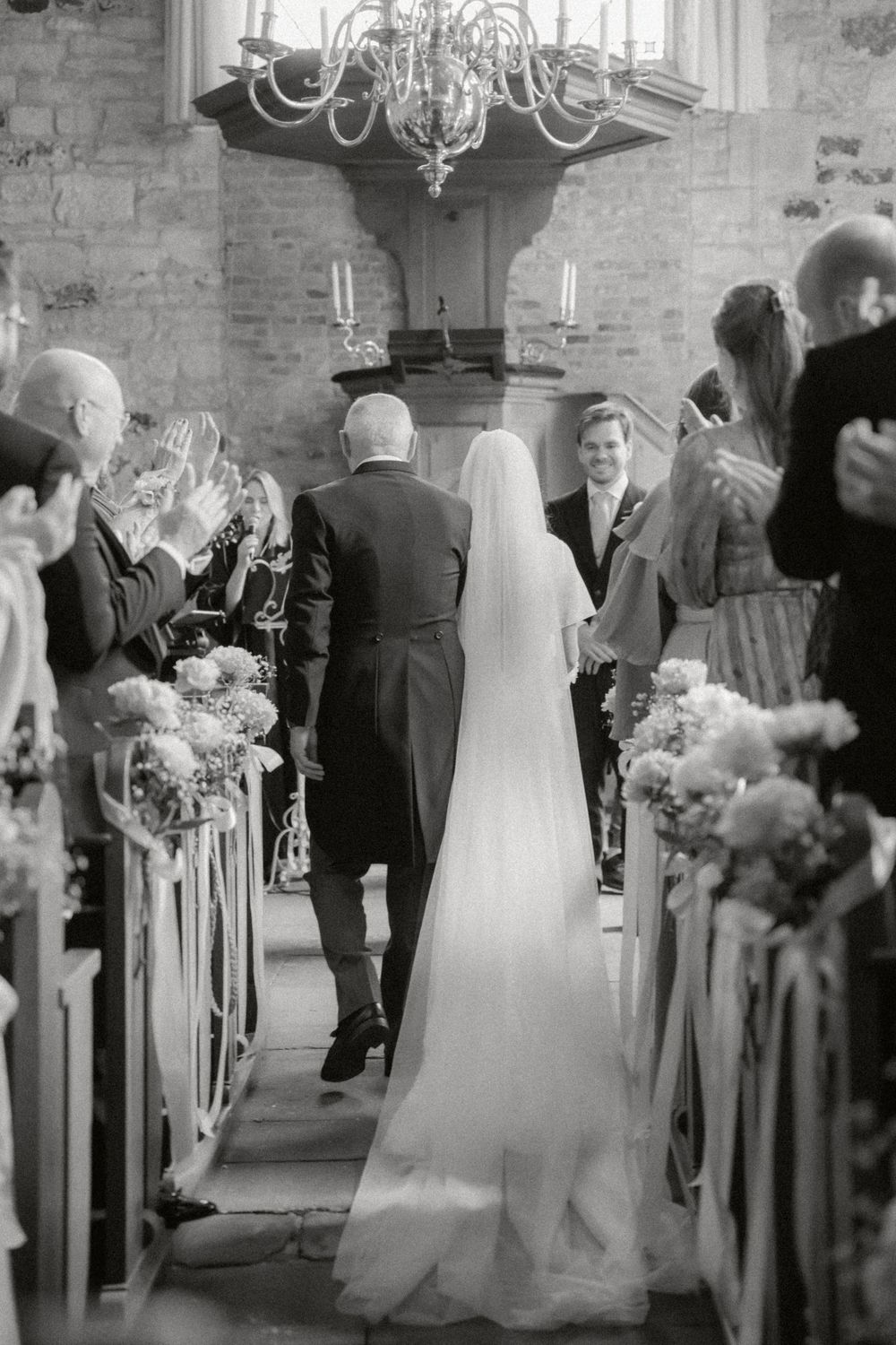 Black and white photo of a wedding ceremony in a church with stone walls and floral decorations along the aisle.