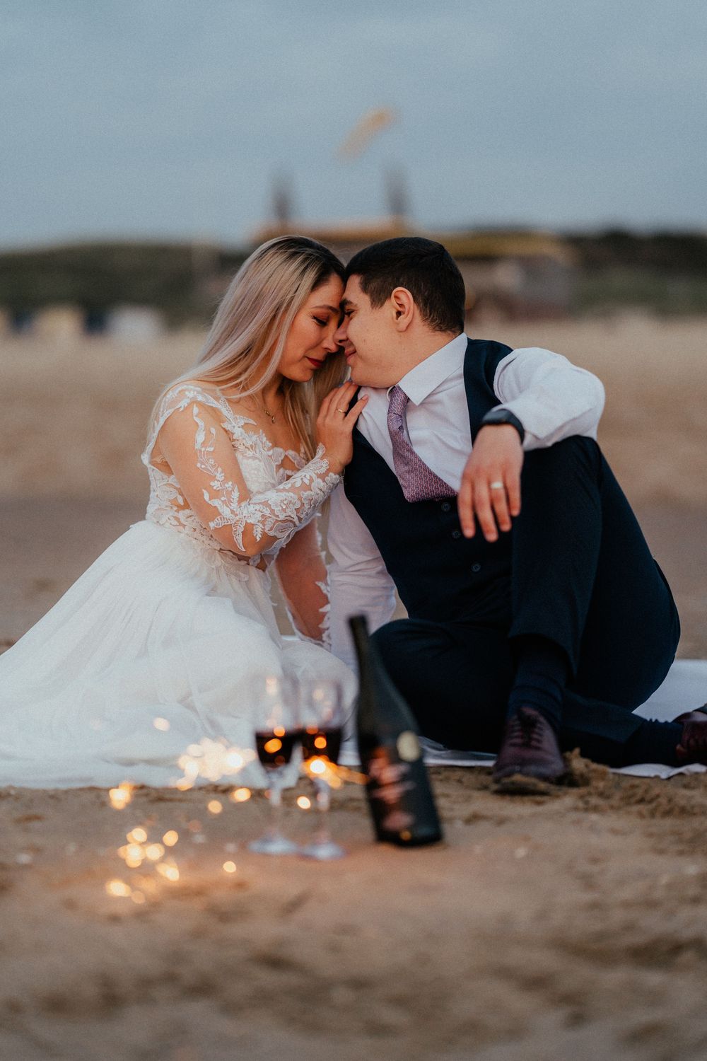 A romantic beach wedding photo with sparklers at sunset showing a couple sitting close together on the sand.