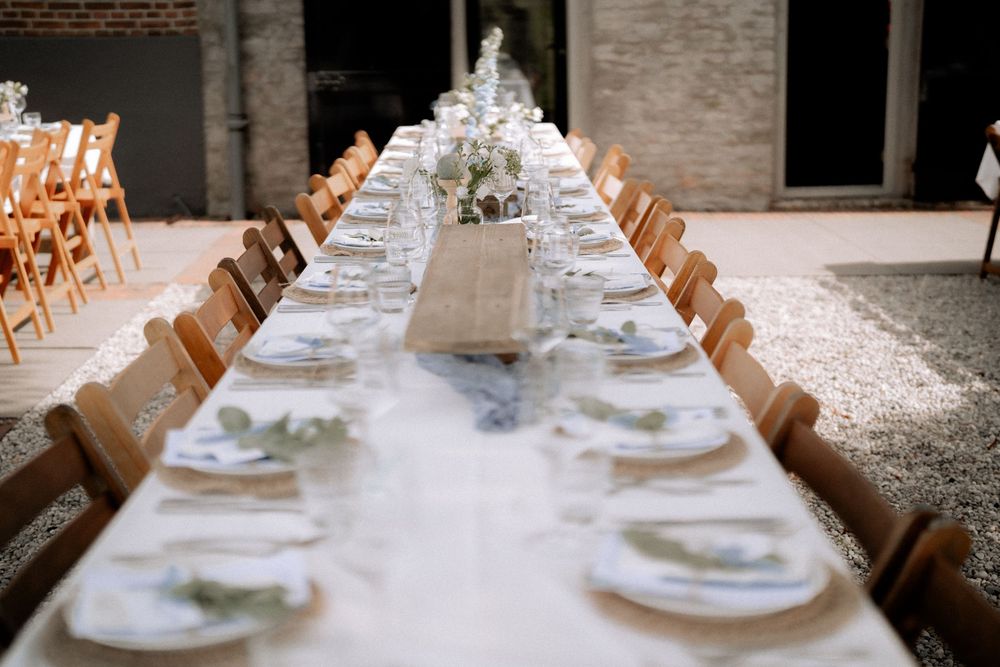 An elegant long wedding reception table with white linens, wooden chairs, and delicate table settings in a rustic venue.