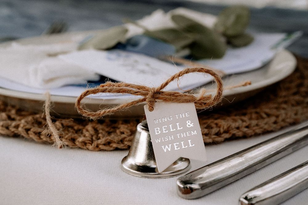 Small silver bell with twine bow and 'Ring if you need wine' tag sits on rustic place setting with plates and cutlery.