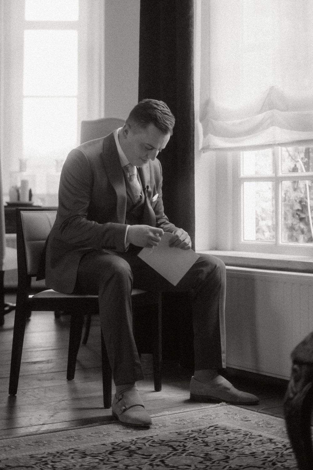 A man in a suit sits by a window reading papers in this vintage black and white photograph from the early 20th century.