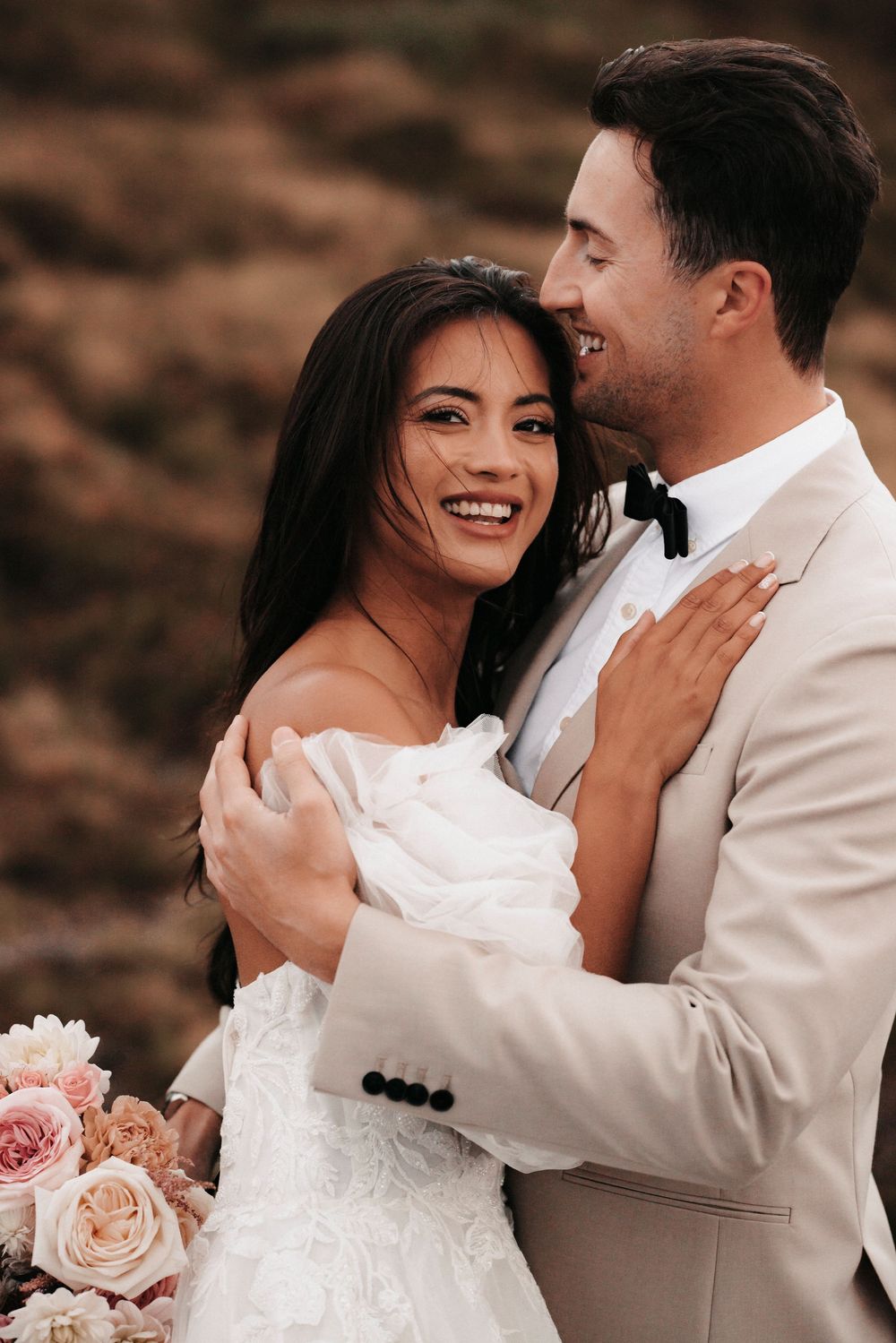 A romantic wedding portrait shows a bride and groom embracing and laughing against a rustic outdoor backdrop with blush roses.