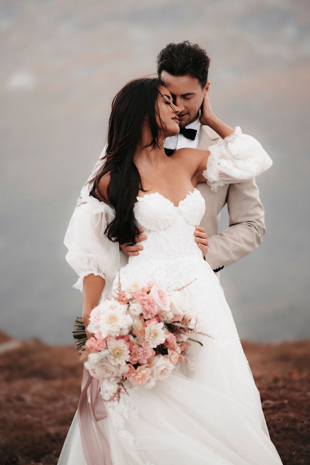 A romantic wedding couple embraces on a hilltop, the bride holding a pink and white bouquet.