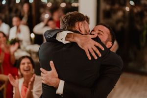 Emotional Wedding photography where the groom shares a warm embrace at a celebration while guests react joyfully in the background.