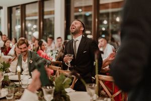 A wedding reception groom laughs joyfully while holding a wine glass during a dinner toast at an elegant indoor dinner