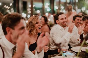 Wedding guests laugh and clap during reception celebration with bokeh lights twinkling in background.