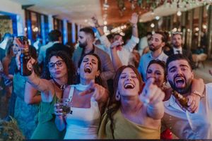 Excited crowd of people cheering and celebrating at a party while holding drinks and raising their hands.