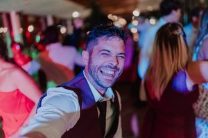 A smiling guest in a dress vest enjoys the wedding party at an indoor reception with colorful lighting in the background.