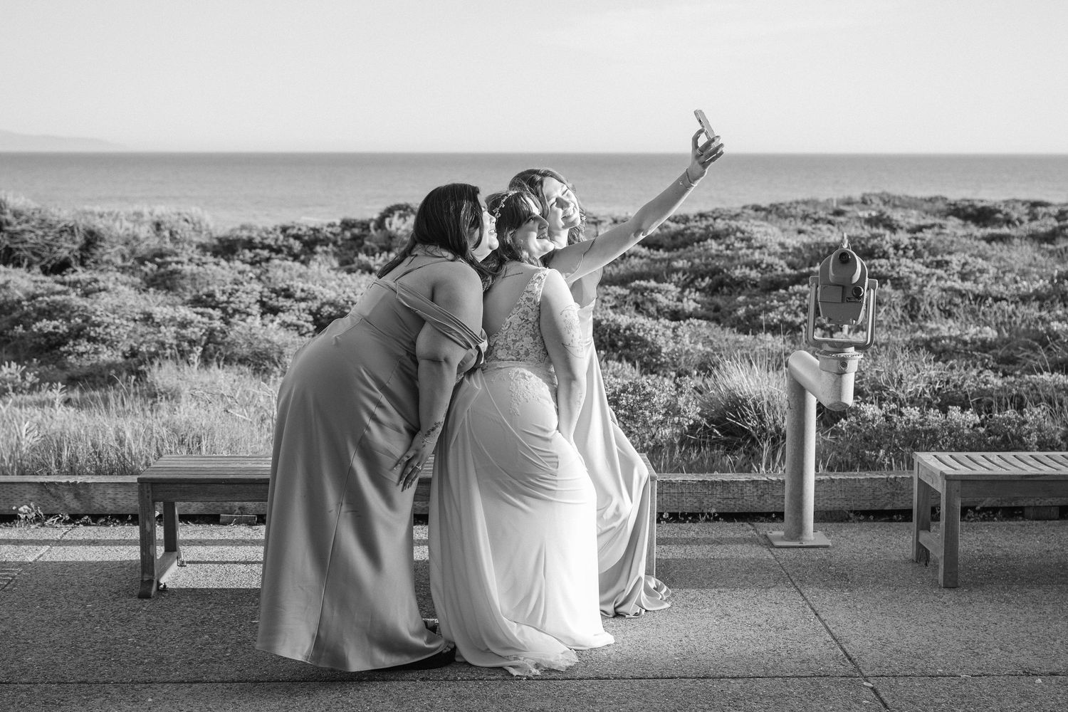Two people in long gowns pose together while taking a selfie on an outdoor terrace overlooking a scenic landscape.