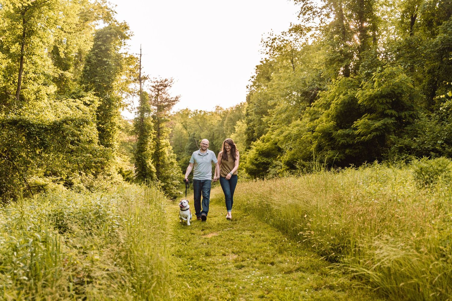 Couple walking with their small dog along a peaceful nature trail surrounded by lush summer greenery.