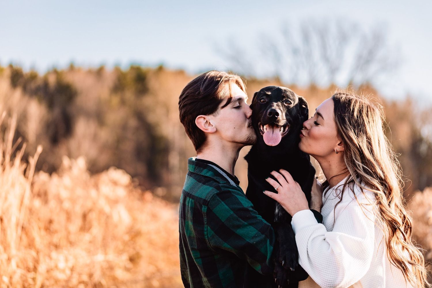 A playful outdoor photo with people kissing a black dog from both sides in a golden autumn field at sunset.