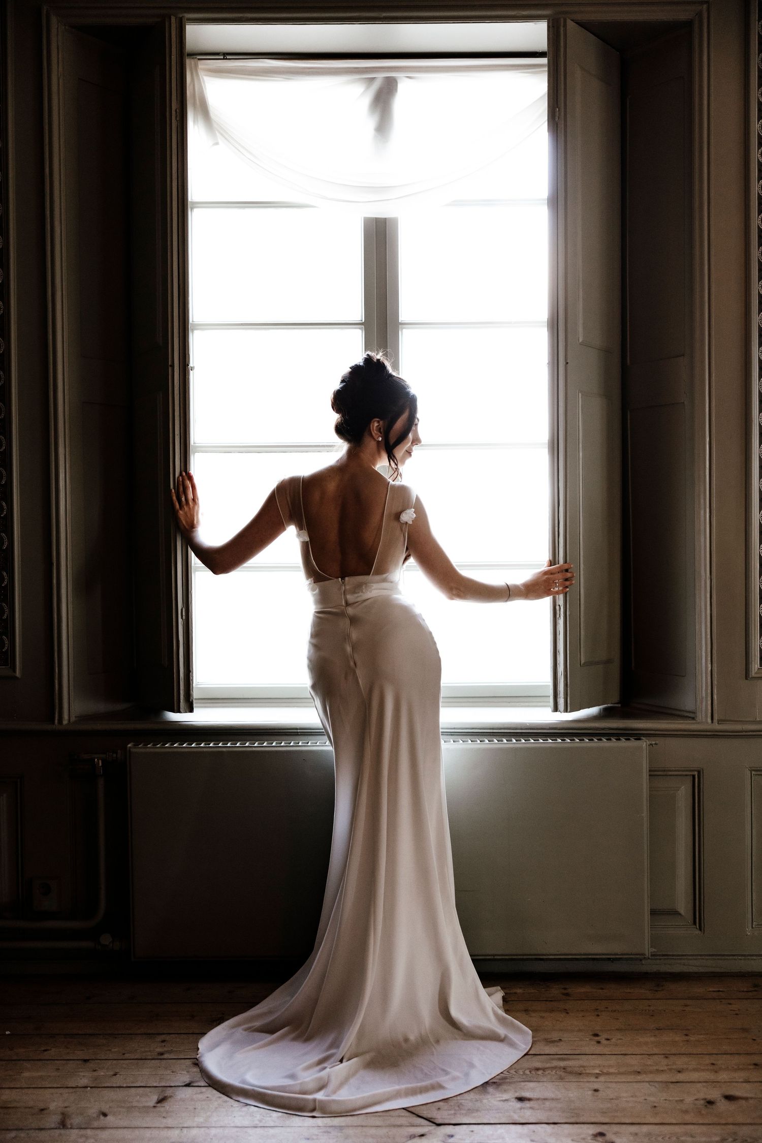 A bride in an elegant white wedding dress stands silhouetted against a bright window in a vintage room.