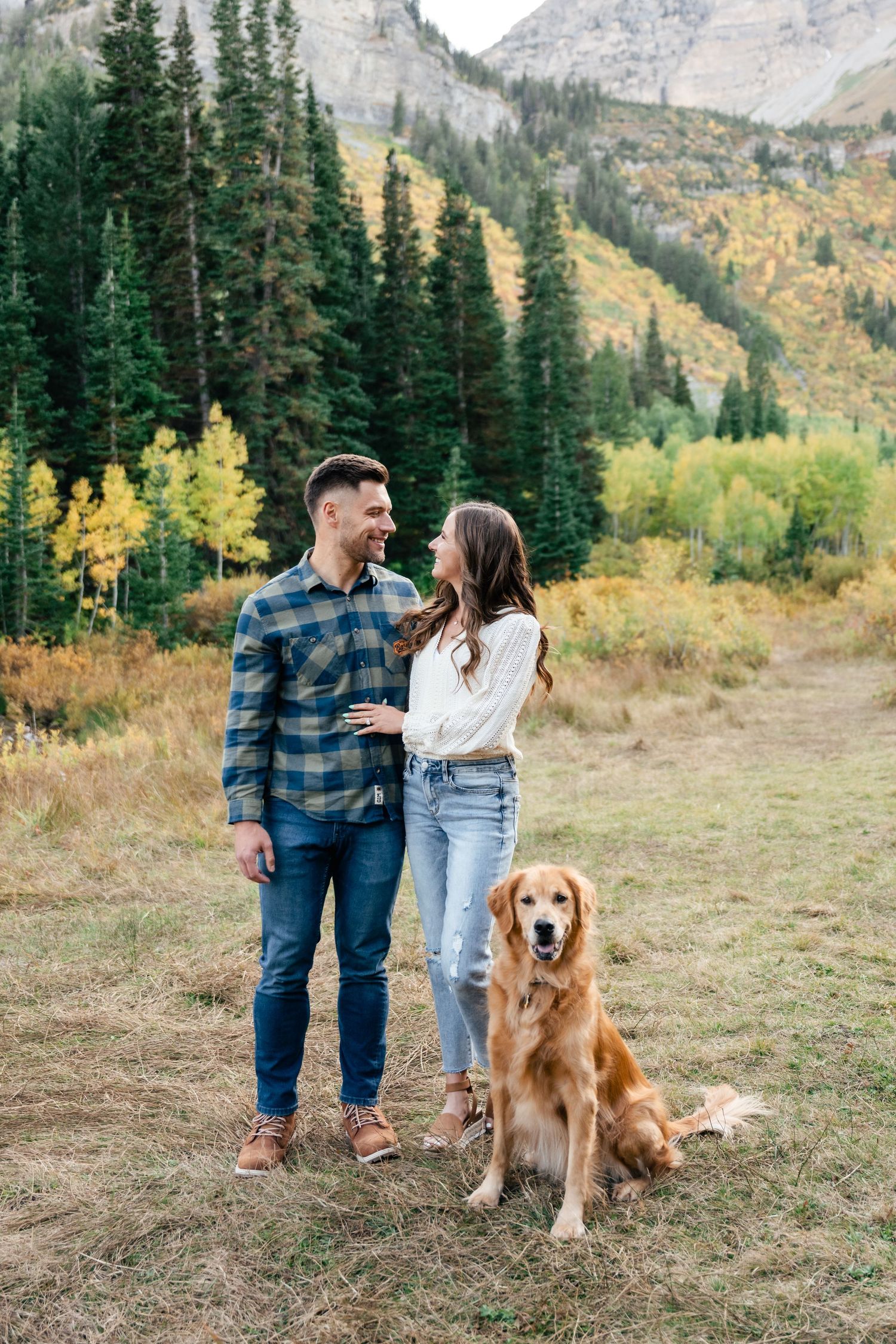 A couple stands on a mountain trail with their Golden Retriever against a backdrop of evergreen trees and autumn colors.