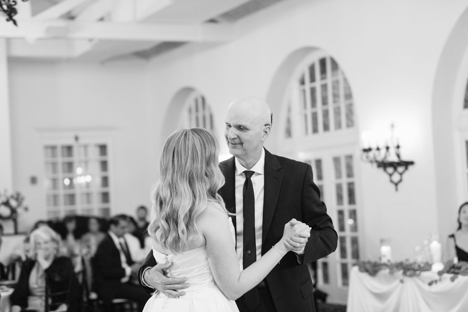 A black and white photo shows a couple dancing in a dip pose while wedding guests watch from their seats in an elegant ballroom.