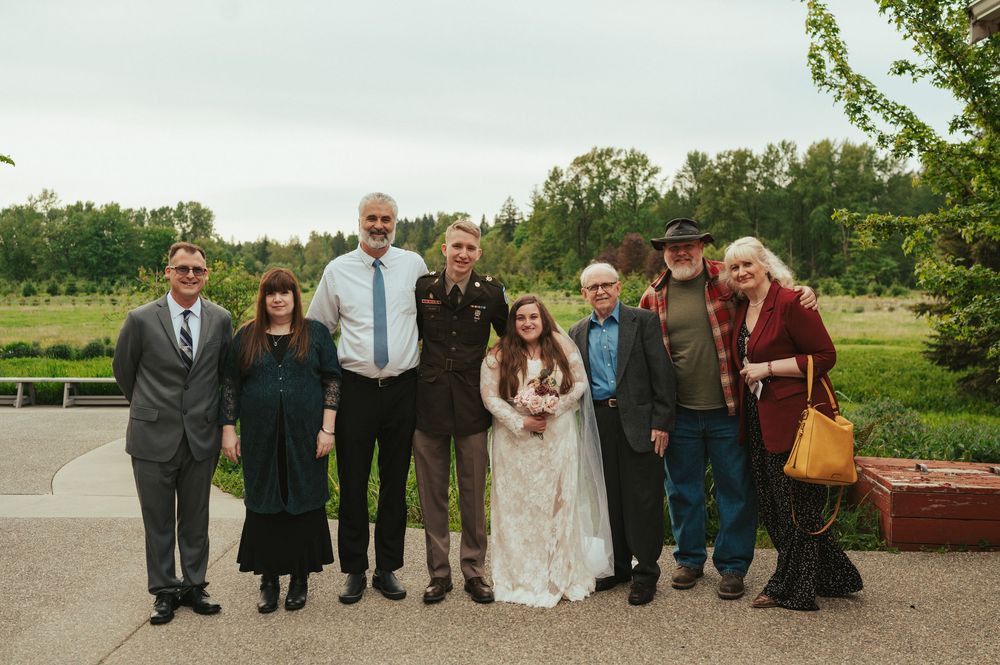 A group of wedding attendees pose outdoors on a sunny day with trees and greenery visible in the background.